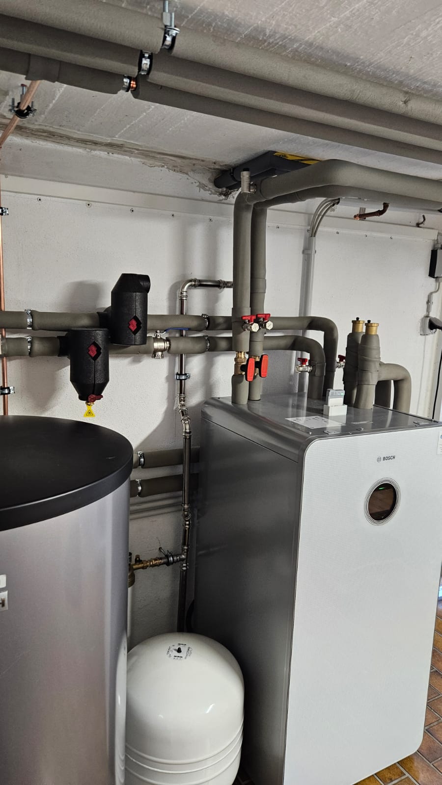 Im Technikraum steht eine Bosch-Wärmepumpe mit integriertem Display, angeschlossen an isolierte Rohrleitungen und einen Pufferspeicher.